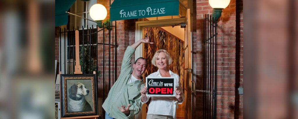 man and woman holding a sign that reads "come in we're open" outside of the Frame to Please store