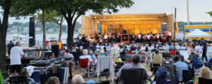 A crowd of people sitting in lawn chairs listening to an orchestra on stage in front of a rive