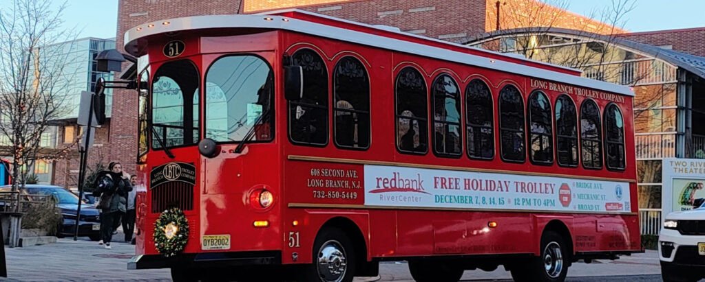 Red trolley with wreath on the front of the vehicle and a banner along the side that reads "Free Holiday Trolley"