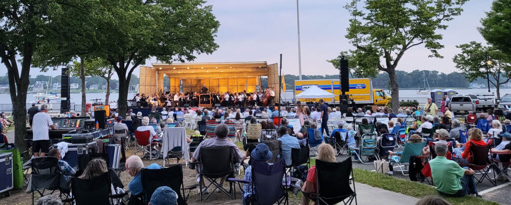 A crowd of people sitting in lawn chairs listening to an orchestra on stage in front of a rive